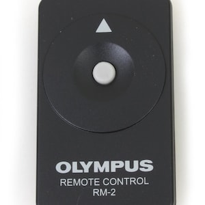 May include: Black Olympus RM-2 remote control with a circular button and a white triangle. The remote has the brand name and model number printed in white. The device is rectangular and has rounded corners.