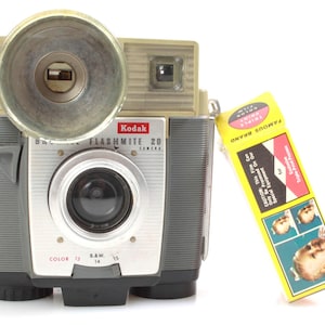 May include: A vintage Kodak Flashmite 20 camera with a built-in flash. The camera is gray and silver with a black lens. The camera has a dial for color and black and white film.