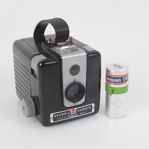 May include: A vintage black Brownie Hawkeye Flash camera with a silver face and a black leather handle. A silver film canister with green and red accents sits next to the camera. The camera has a rectangular shape with a lens and flash.