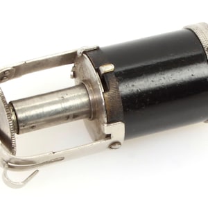 Op de afbeelding: Een zilveren en zwarte metalen filmwikkelapparaat met een draaiknop en een zwarte cilinder. Het apparaat wordt gebruikt om film op een spoel te wikkelen.