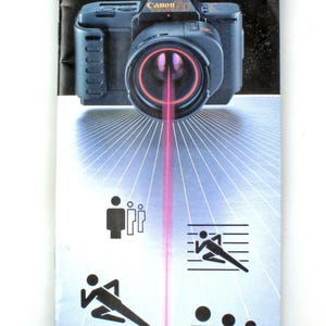 Puede incluir: Una cámara Canon T80 negra con un rayo láser rojo apuntando hacia abajo. La cámara tiene el texto "Canon T80" en la parte delantera. El texto "Auto Focus System Picture Selector" está impreso debajo de la cámara. La imagen tiene cuatro figuras de palo negras en diferentes poses.