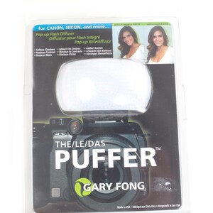 May include: A pop-up flash diffuser for Canon, Nikon, and more. The product is called "The Puffer" by Gary Fong. The packaging is black and white, with a clear plastic front. The diffuser is white and oval-shaped.
