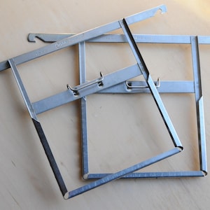 May include: Two silver metal picture frames with a hook on each corner. The frames are stacked on top of each other.