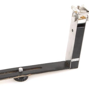 Puede incluir: Un soporte de flash de cámara vintage, con un diseño en ángulo recto. El soporte es de metal con un acabado negro y plateado. Tiene un tornillo de montaje y una zapata para flash. La marca "ROWI" es visible.