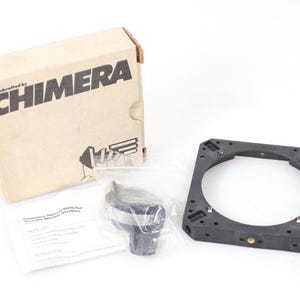 Puede incluir: Un soporte de montaje negro para modificador de luz Chimera con un anillo de velocidad Chimera plateado y negro. El soporte es de plástico y tiene forma cuadrada. El anillo de velocidad es de metal y tiene forma redonda. El soporte y el anillo de velocidad están ambos en una caja blanca con la palabra "CHIMERA" impresa en ella.