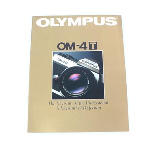 May include: A brochure for the Olympus OM-4Ti camera. The brochure has a gold background with the word "OLYMPUS" at the top. The image in the center shows a close-up of the camera. The text at the bottom reads "The Measure of the Professional. A Measure of Perfection."