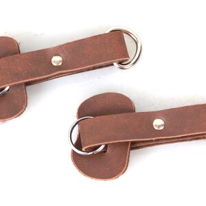 May include: Two brown leather straps with metal rings and a silver rivet. The straps are designed to be used as a closure system for bags or other leather goods.