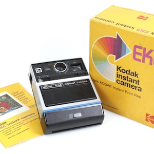 Puede incluir: Una cámara instantánea Kodak EK6 vintage con un cuerpo negro y plateado, una caja amarilla y un manual. La caja presenta un diseño de flecha colorida y el texto "Kodak EK6 instant camera". El manual tiene una foto de una persona.