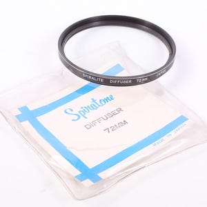 May include: A black Spiratone Diffuser 72mm lens filter with text that reads "SPIRALITE DIFFUSER 72 mm JAPAN". The filter is on top of a clear plastic bag with a white label and blue accents. The label reads "Spiratone DIFFUSER 72MM".