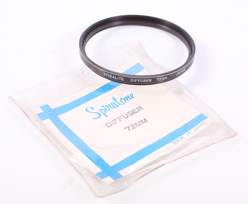 Peut inclure: Un filtre d'objectif Spiratone Diffuser noir de 72 mm avec le texte "SPIRALITE DIFFUSER 72 mm JAPAN". Le filtre est pos&eacute; sur un sac en plastique transparent avec une &eacute;tiquette blanche et des accents bleus. L'&eacute;tiquette indique "Spiratone DIFFUSER 72MM".