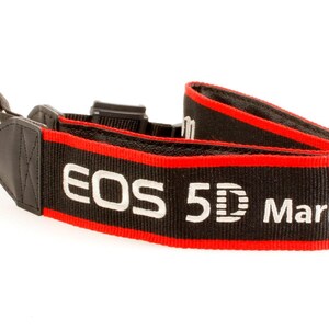 May include: A black camera neck strap with red trim and white embroidered text that reads EOS 5D Mark III. The strap features black leather-like end tabs with black plastic adjustment buckles and attachment loops for securing to a camera body.