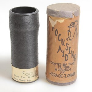 May include: A black cylindrical object with a textured surface next to a brown cylindrical container. The container has the words "FOCUSING AIDING" and "CRAFTED BY HAND IN THE WORKSHOP OF HORACE J. CHASE" written on it.
