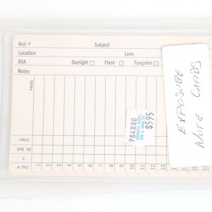 May include: A clear plastic sleeve containing a beige exposure note card. The card has a grid for recording data, with the words "EXPOSURE NOTE CARDS" written on a white label. A sticker with a price is visible.