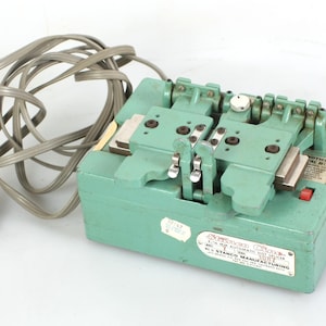 Könnte beinhalten: Ein grüner Drahtabisolierer aus Metall mit einem grauen Netzkabel und Stecker. Die Maschine hat mehrere Komponenten, darunter Hebel, Schrauben und einen roten Knopf. Ein Etikett trägt die Aufschrift "STANCO MANUFACTURING".
