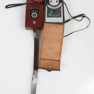 May include: A vintage light meter with a leather case. The meter is black and silver with a circular dial and a needle. The case is burgundy leather with a long strap. The meter is designed for photography.