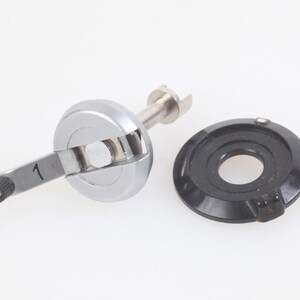 May include: A silver-colored sewing machine part with a black dial and a lever marked with the number 1. The part is designed for a sewing machine and is set against a white background.