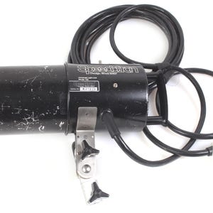 Puede incluir: Cabezal de flash Speedotron Speedtorch negro con un cable de alimentación negro enrollado y un soporte de montaje plateado. El cabezal del flash tiene el logotipo de Speedotron y la información del modelo.