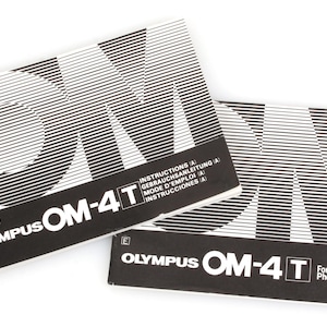 May include: Two black and white Olympus OM-4T camera instruction manuals. The manuals feature the text "OM-4T" in white, with the Olympus logo and the text "For Your Creative Photography (B)". The covers have a geometric design.