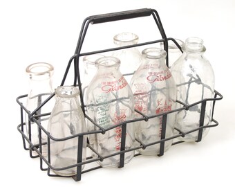 May include: A black metal milk bottle carrier with a handle holds six glass milk bottles. The bottles are clear and have red and green labels with the text "For Delivery Only Glendora Dairy".