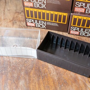 May include: Two empty storage cases for audio cassette tapes. One case is clear plastic and the other is brown with slots for the tapes. The cases are on a wooden surface.