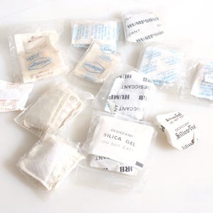 May include: A collection of small white silica gel packets with the text "Desiccant Silica Gel Do Not Eat" and "Desiccant Silica-Tre" printed on them. Some packets are in clear plastic bags.