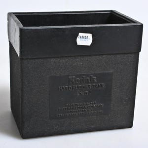 May include: A black Kodak Hard Rubber Tank 4x5. The box has a textured surface and a rectangular shape with an open top. The Kodak logo and product information are embossed on the front. A small white sticker is affixed to the top.