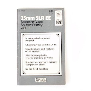 May include: A gray softcover book titled 35mm SLR EE Selection Guide Shutter-Priority Vol. I. The cover features white text listing topics such as choosing your 35mm SLR EE, specifications, features, shutter-priority systems, and comparison charts.