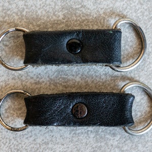 以下が含まれることがあります： 銀色の金属製のリングが付いた黒い革のストラップが2本。各ストラップには黒いボタンが付いています。