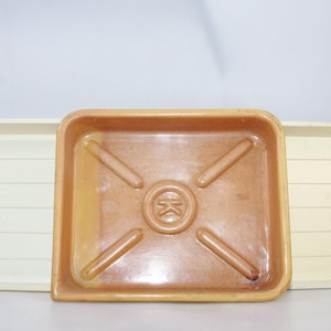 May include: Three rectangular trays. A square, light brown tray with rounded corners and a central embossed design is in the center. Two off-white trays with a slatted design are on either side. The trays are likely for gardening or crafts.