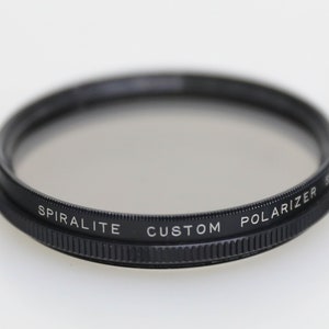 Może przedstawiać: Czarny, okrągły filtr do obiektywu aparatu z nadrukiem "SPIRALITE CUSTOM POLARIZER" na krawędzi.