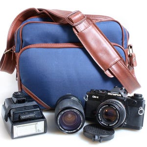 May include: A blue camera bag with brown leather trim and strap, alongside a black Olympus OM-4 camera, a lens, and a flash unit. The bag has a rectangular shape and a shoulder strap.