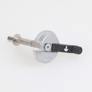 Puede incluir: Un abridor de botellas de vino plateado con una palanca negra y un pequeño mango negro redondo. El abridor tiene una varilla metálica y una base circular. Una flecha blanca indica la dirección de uso.