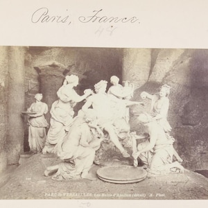 May include: A sepia-toned photograph depicting the sculpture group Les Bains d'Apollon in Versailles, France. The image shows multiple figures surrounding a central seated figure, with text at the top reading Paris, France and at the bottom PARC de VERSAILLES, Les Bains d'Apollon (détail) X- Phot.