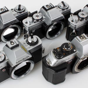Puede incluir: Una colección de cámaras de película Minolta y Konica vintage. Las cámaras tienen una combinación de colores plateado y negro, con lentes y diales visibles. Las cámaras Minolta están etiquetadas como "XG-1". La Konica está etiquetada como "FS-1".