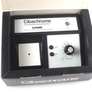 以下が含まれることがあります： Ilford製の白黒Cibachrome Exposure Monitor M-1。本体には、黒いキャリブレーションノブ、HighとLowとラベル付けされた2つの赤いインジケーターライト、電源用のトグルスイッチを備えたシルバーのコントロールパネルがあります。デバイスは、前面に製品名が印刷された黒い箱に収められています。
