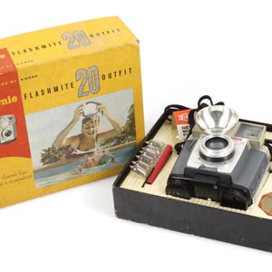 May include: Vintage Kodak Brownie Flashmite 20 Outfit camera set. The camera is gray and silver, with a flash attachment. The set includes the camera, flash, and accessories, all in a yellow and red box.