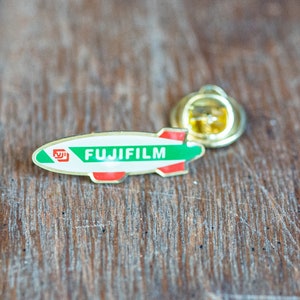 May include: A gold-colored pin shaped like a blimp with the words "FUJIFILM" printed in black on a white background. The blimp has red and green accents.