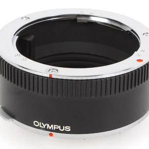 Könnte beinhalten: Ein schwarzer und silberner Olympus-Objektivadapter. Der Adapter hat einen geriffelten schwarzen Körper mit einem silbernen Ring an der Oberseite. Das Wort "OLYMPUS" ist weiß auf der Seite aufgedruckt. Rote Markierungen sind sichtbar.