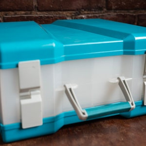 Può includere: Una scatola di plastica blu e bianca con una maniglia e chiusure. La scatola è chiusa e ha un interno bianco.