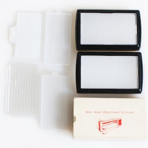 May include: A collection of camera accessories, including two black-framed light diffusers, several clear plastic diffusers, and a small rectangular box. The box reads "Wide Angle Attachment Enclosed" in red text. The items are arranged on a white surface.