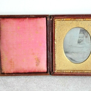 May include: A vintage hinged case with a dark brown exterior. The left side features a padded pink fabric interior. The right side contains a black and white oval portrait of a person wrapped in fabric, framed by an ornate gold-colored metal border.