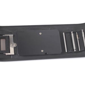 May include: A black rectangular camera back component featuring a central flat plate, a rectangular foam-lined opening on the left side, and a series of three vertical metallic pins secured on the right side within a molded plastic frame.