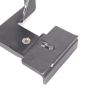 May include: A black metal bracket with a rectangular base and an L-shaped arm. The bracket has a rubber pad and a screw for attachment. The item is likely a camera accessory or mounting component.
