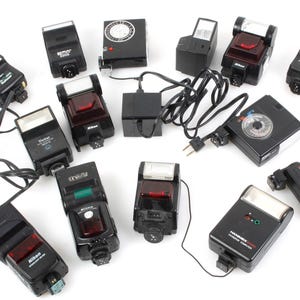 Op de afbeelding: Een verzameling van 12 zwarte flitser-eenheden voor camera's. De flitser-eenheden zijn van verschillende groottes en vormen, en sommige hebben een rood of groen lampje.