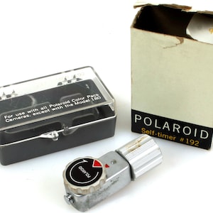 Op de afbeelding: Een vintage Polaroid zelfontspanner, model #192, met een zilverkleurige metalen behuizing en een zwart-rode wijzerplaat. De afbeelding toont ook een zwarte plastic doos met een doorzichtig deksel en een kartonnen doos met het Polaroid logo en tekst. De tekst op de behuizing luidt: "Voor gebruik met alle Polaroid Color Pack Camera's, behalve de Model 180."