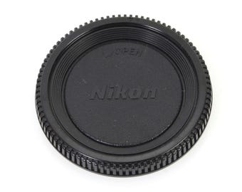 May include: Black Nikon lens cap with a ridged edge and the word "Nikon" printed in white.