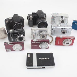 Puede incluir: Una colección de cámaras digitales en varios colores, incluyendo negro, plateado, rojo y azul. Las cámaras son de diferentes tamaños y marcas, como Nikon, Canon y Polaroid. La imagen está tomada desde un ángulo alto.