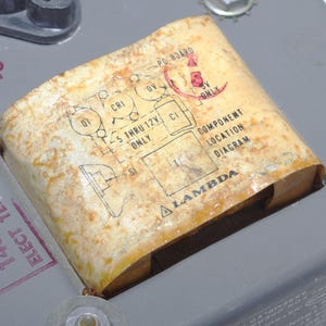 以下が含まれることがあります： 色あせた、古びた外観の電子部品のクローズアップ。 この部品には、「Lambda」や「Component Location Diagram」などの図とテキストが記載された紙ラベルが付いています。 背景は、ボルトが付いた灰色の金属面です。