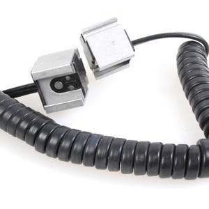 Könnte beinhalten: Ein schwarzes Spiralkabel mit zwei silbernen Steckern. Die Stecker sind quadratisch und mit einem kleinen schwarzen Kabel an das Kabel befestigt.