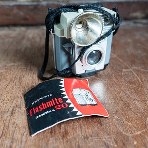 May include: A vintage Brownie Flashmite 20 camera with a black strap and a flash attachment. The camera is silver and black and has a red and black instruction booklet.
